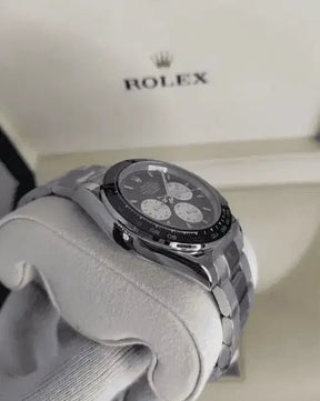 A Rolex Daytona wristwatch with a black and white chronograph dial rests on a soft white cushion, its silver finish gleaming, while the signature Rolex box sits in the background—an iconic luxury timepiece for collectors.