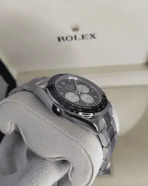 A Rolex Daytona wristwatch with a black and white chronograph dial rests on a soft white cushion, its silver finish gleaming, while the signature Rolex box sits in the background—an iconic luxury timepiece for collectors.