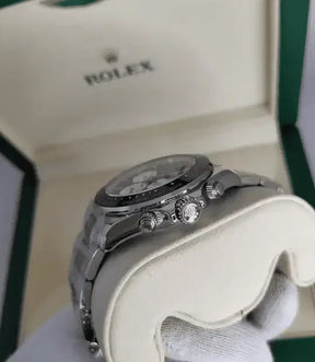 A close-up of a silver Rolex Daytona held in a gloved hand, with a green and cream Rolex box in the background, highlights this iconic stainless steel luxury watch.