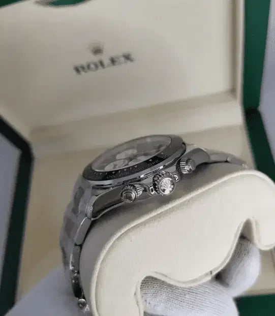 A close-up of a silver Rolex Daytona held in a gloved hand, with a green and cream Rolex box in the background, highlights this iconic stainless steel luxury watch.
