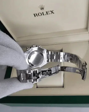 A gloved hand holds a Rolex Daytona, displaying its metal clasp and stainless steel case back above an open Rolex box with the logo visible inside the lid.