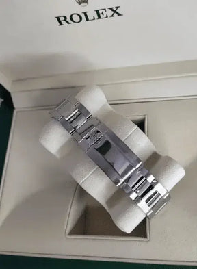 A Rolex Daytona luxury watch with a metallic stainless steel bracelet is showcased in a cream Rolex case, resting on a padded cushion. The display highlights the clasp and band; the watch face is not visible.