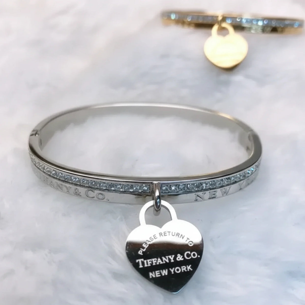 A silver Bracelete Bangle Return to Tiffany with a heart-shaped "Please return to Tiffany & Co. New York" charm rests on a white, fluffy surface; in the softly blurred background, a matching bracelet completes the luxurious scene.