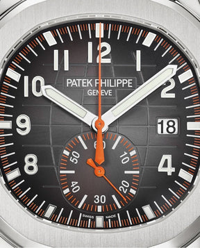 Close-up do Relógio Patek Aquanaut com mostrador preto, números brancos em destaque, ponteiros brancos e laranja, janela de data às 3h e subdial de segundos às 6h—um verdadeiro relógio de luxo com detalhes sofisticados.